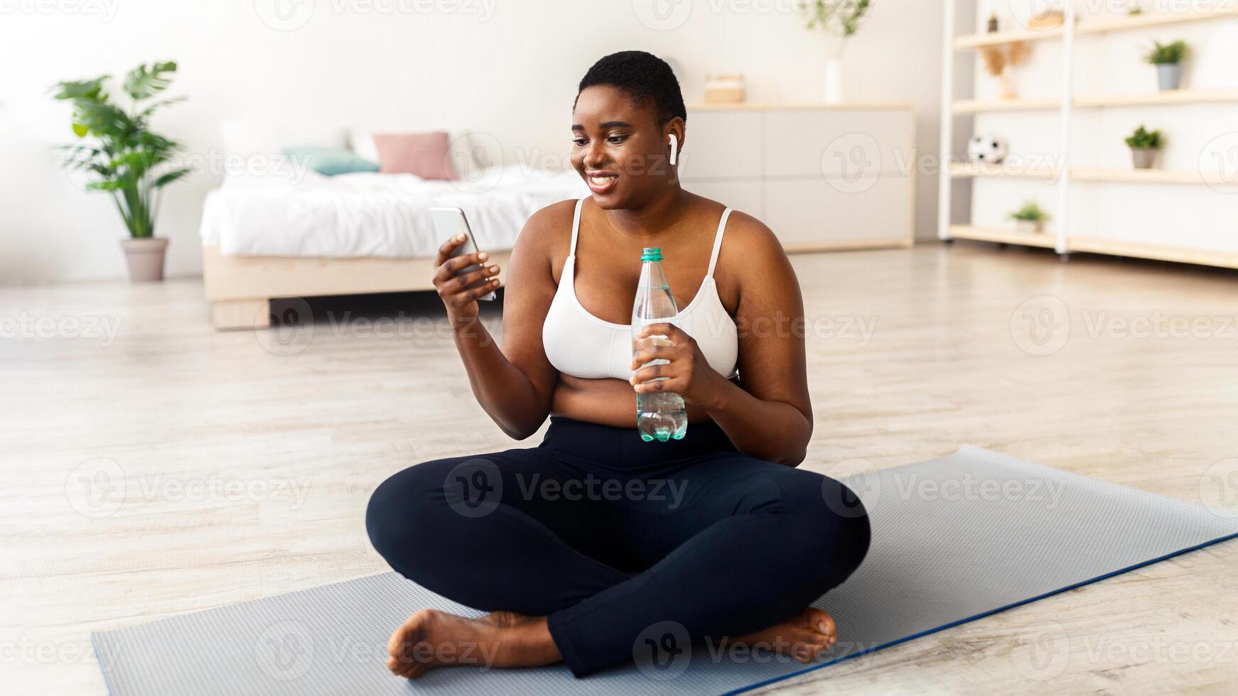 Plus size young black woman with bottle of water and cellphone surfing web on break from home training, checking music online. Curvy African American female using mobile phone after workout photo