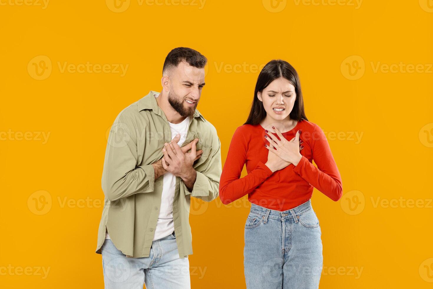 A young European couple stands close together, both placing their hands over their chests, conveying deep feelings of love and affection. Their expressions reflect a strong emotional connection. photo