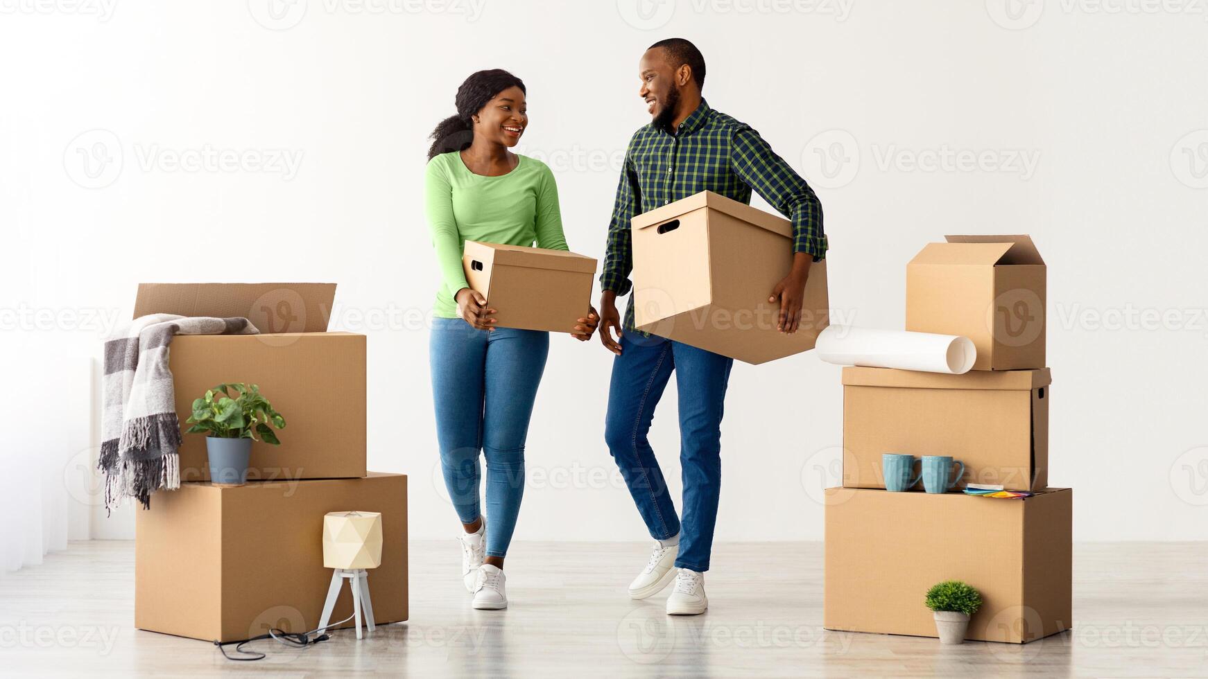Home Relocation Concept. Portrait Of Joyful African American Spouses Walking With Cardboard Boxes After Moving To New Flat, Happy Black Couple Carrying Belongings And Smiling To Each Other photo