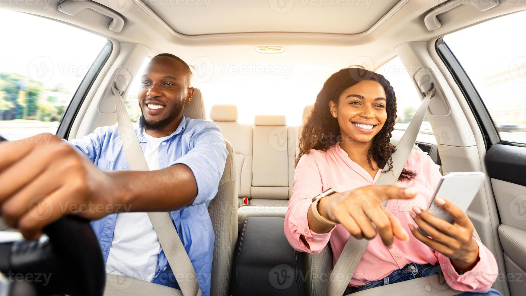 Portrait of smiling African American couple sitting inside luxury car, cheerful man holding steering wheel, woman with mobile phone pointing at the road, showing way, using digital map application photo