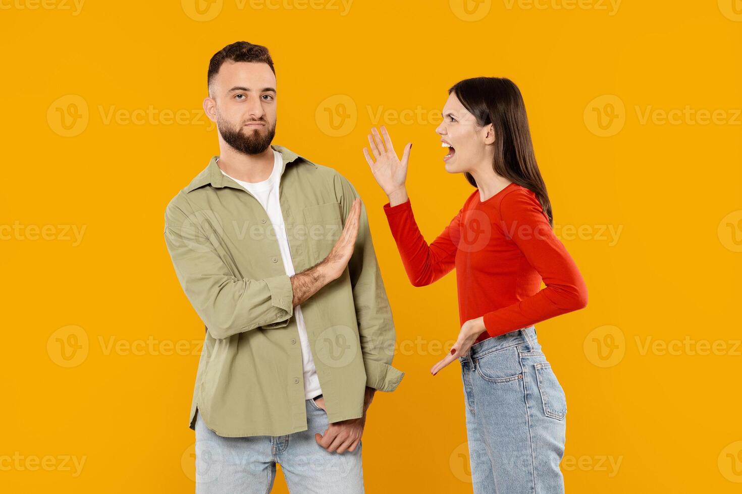 A young couple stands together against a bright yellow background. The woman is animatedly expressing her feelings while the man appears calm, gesturing with a hand to indicate disagreement. photo