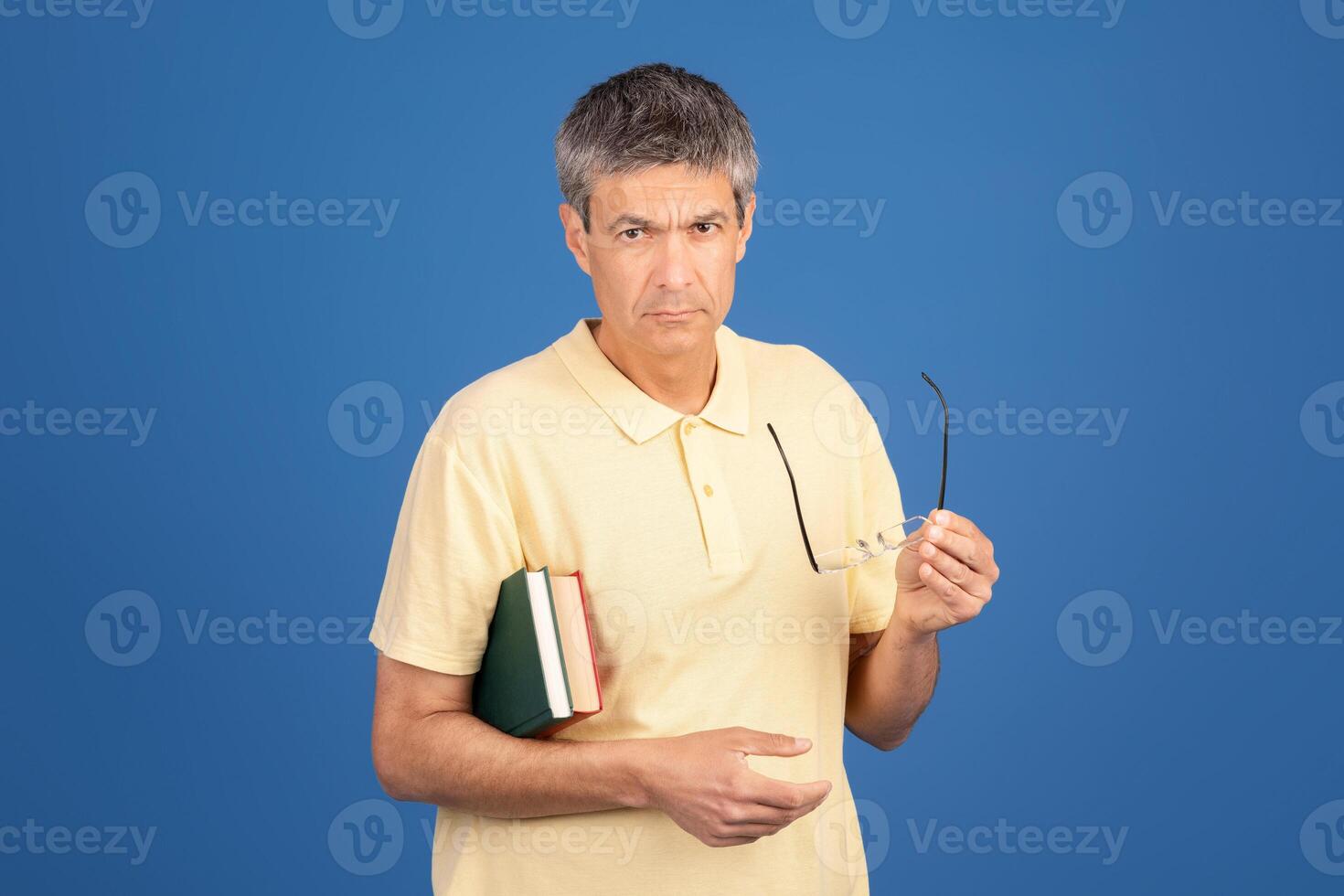 A middle aged man in his 50s stands against a blue background. He holds glasses in one hand and books in the other, looking contemplative and slightly isolated in his surroundings. photo