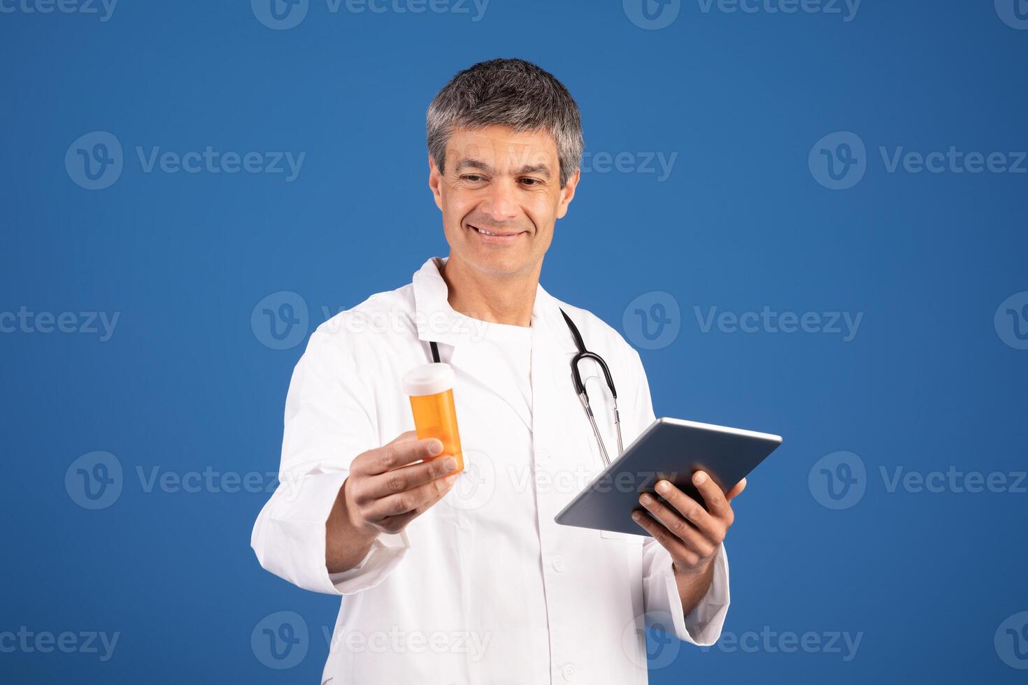 A middle aged European doctor smiles as he examines a prescription bottle in one hand, while holding a tablet in the other. His white coat and stethoscope indicate his medical practice. photo