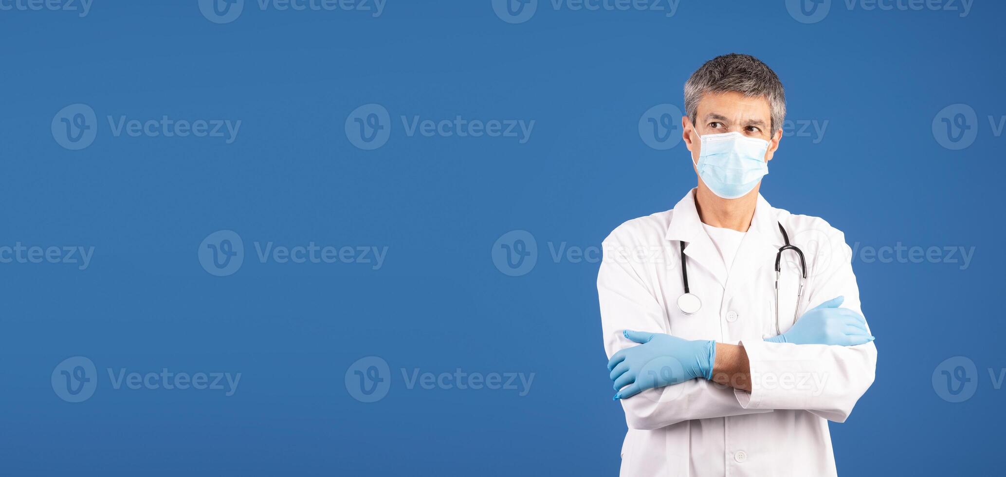 A middle aged Caucasian man dressed in a white lab coat and gloves stands with his arms crossed. He wears a surgical mask, emphasizing his role in healthcare, copy space photo