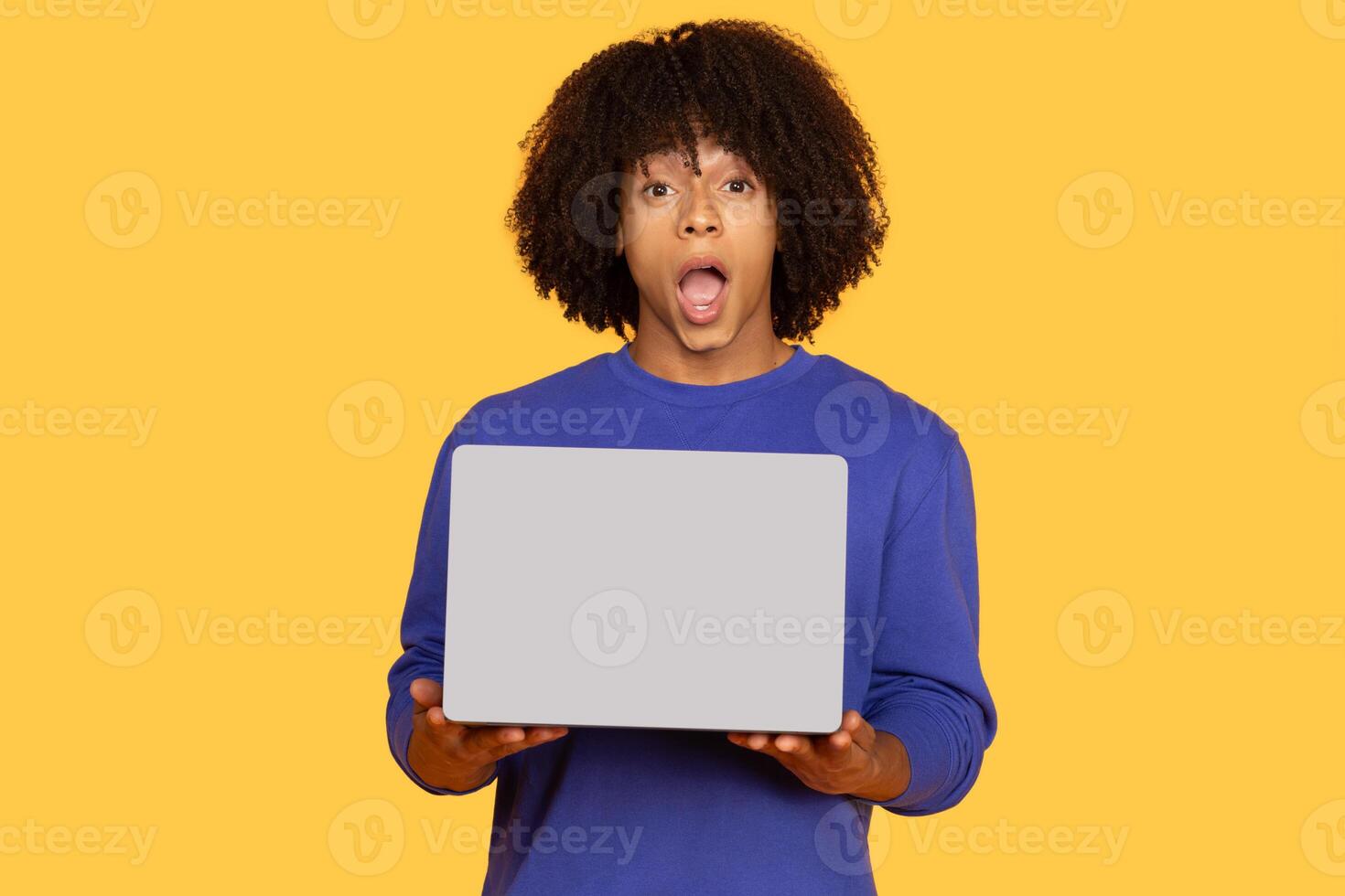 A surprised young man holds a laptop in front of him, showcasing his expressive reaction. He has curly hair and wears a blue sweater, creating a vivid contrast with the yellow background. photo