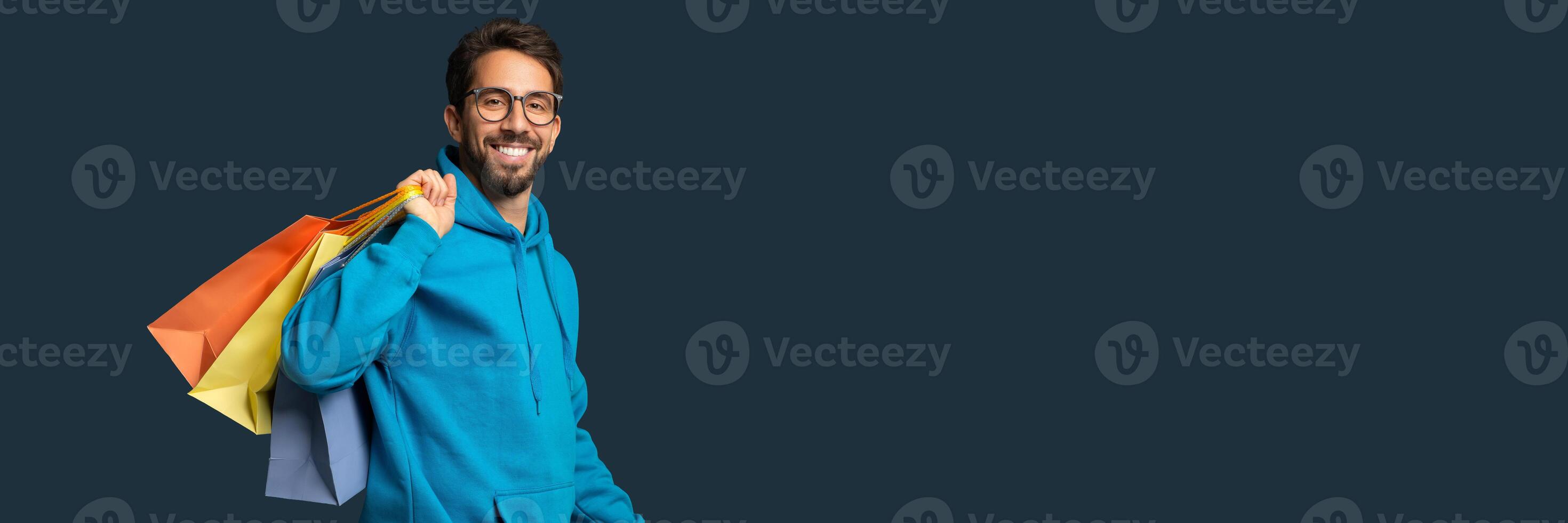 Shopping Concept. Handsome Smiling Young Man Holding Colorful Paper Shopper Bags, Happy Millennial Guy In Eyeglasses Carrying Purchases, Standing Against Blue Studio Background, Free Space photo