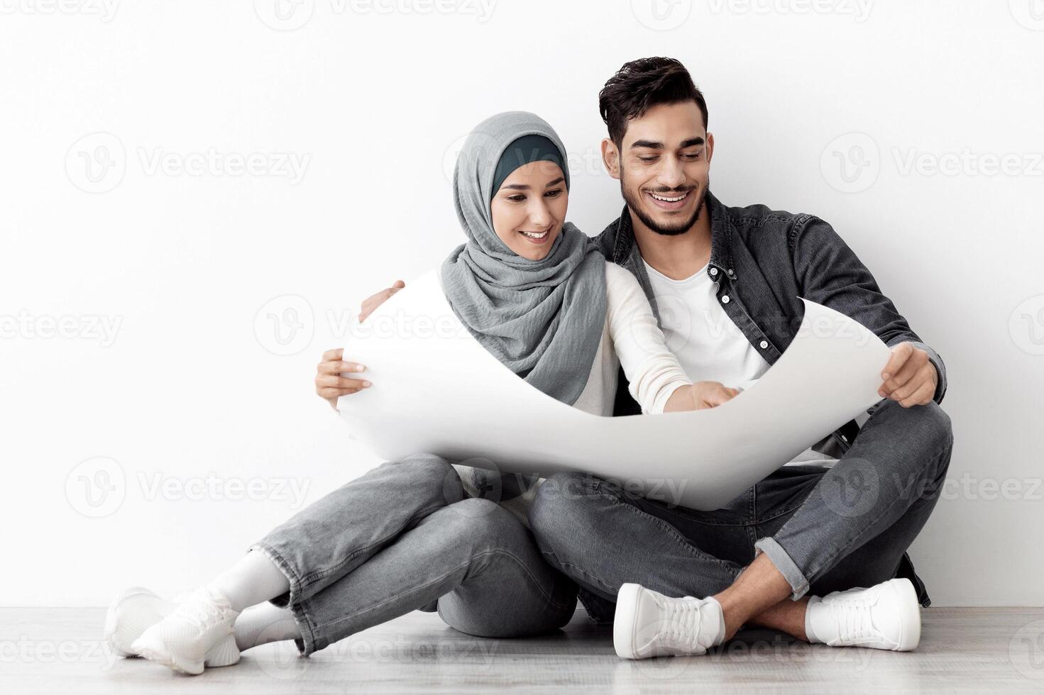 Cheerful middle-eastern family husband and wife in hijab sitting on floor in empty room, holding apartment scheme, copy space for advertising, white blank wall background, panorama photo