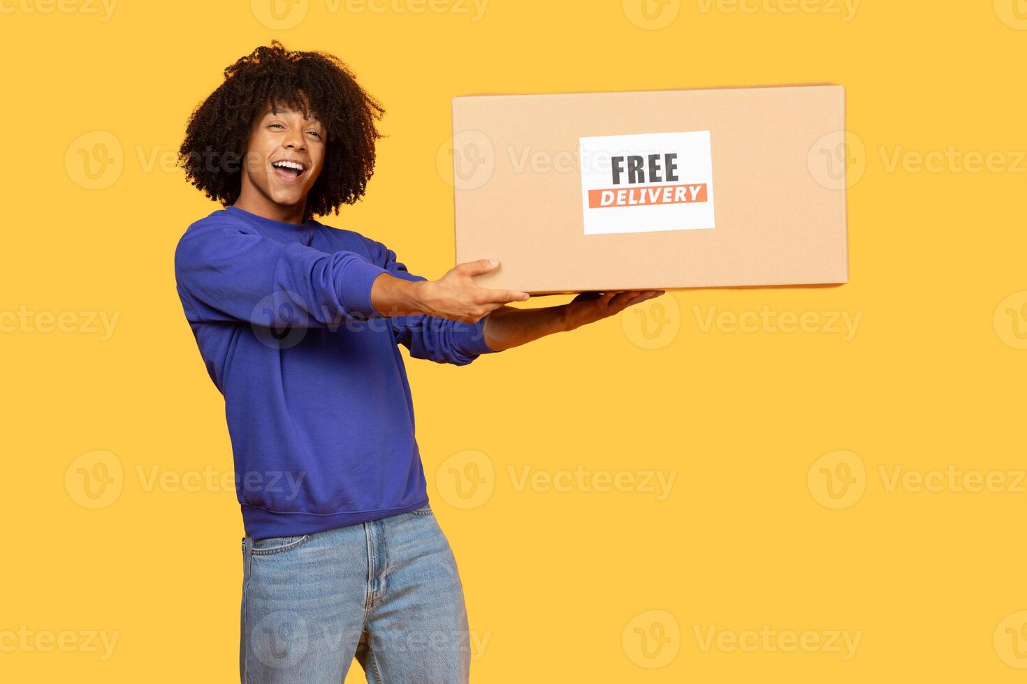 A cheerful young man with curly hair joyfully holds a delivery package that reads free delivery. He wears a blue sweatshirt with a vibrant yellow background, embodying a lively lifestyle. photo