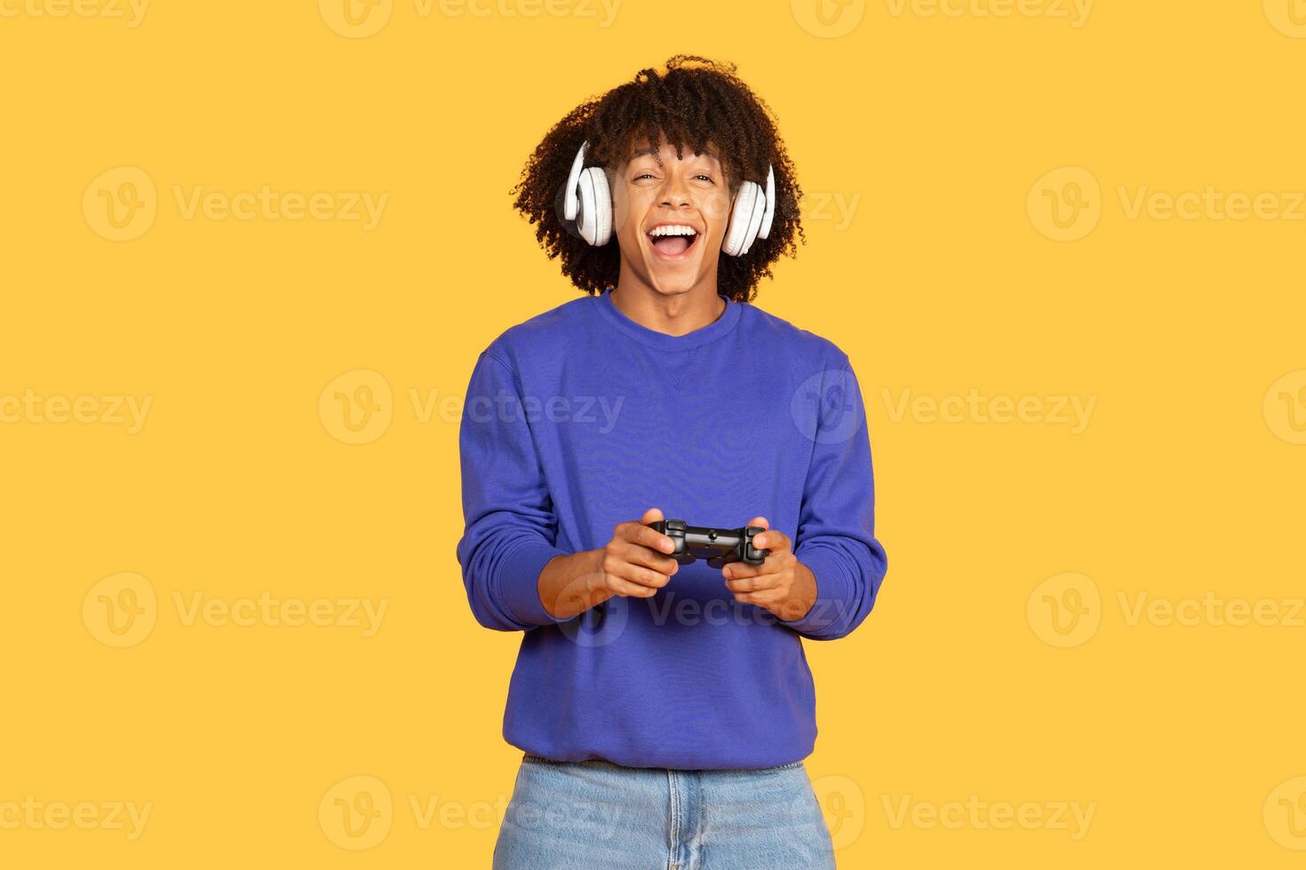 A young black man with curly hair smiles while holding a game controller. He is wearing headphones and is immersed in gaming, showcasing a lively atmosphere in a bright yellow space. photo