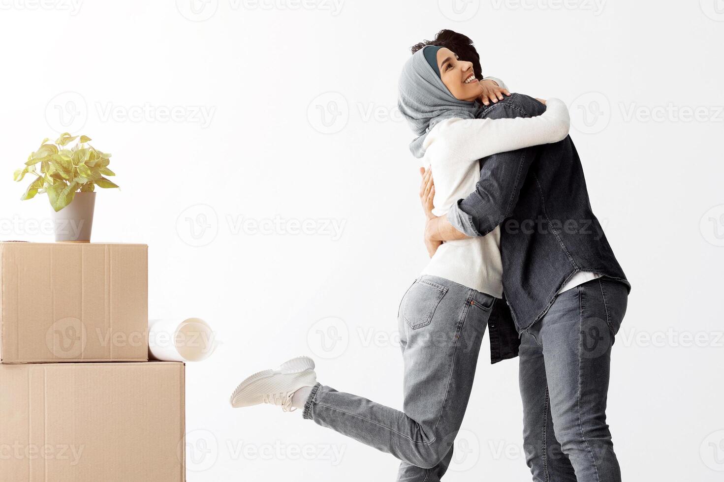 Emotional young muslim lady in hijab hugging her husband, middle-eastern man and woman standing among paper boxes and other stuff in empty room, family moving in new house, full length, copy space photo
