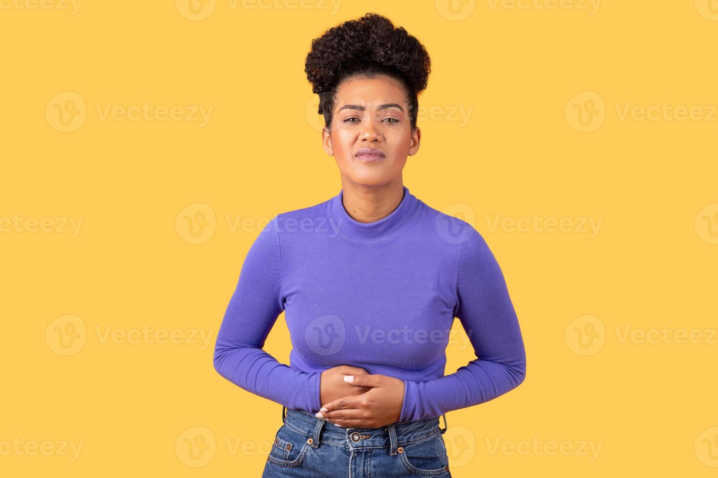 A young Hispanic woman stands alone with her hands on her stomach, showing a thoughtful expression. The bright yellow backdrop adds vibrancy to her relaxed yet serious demeanor. photo
