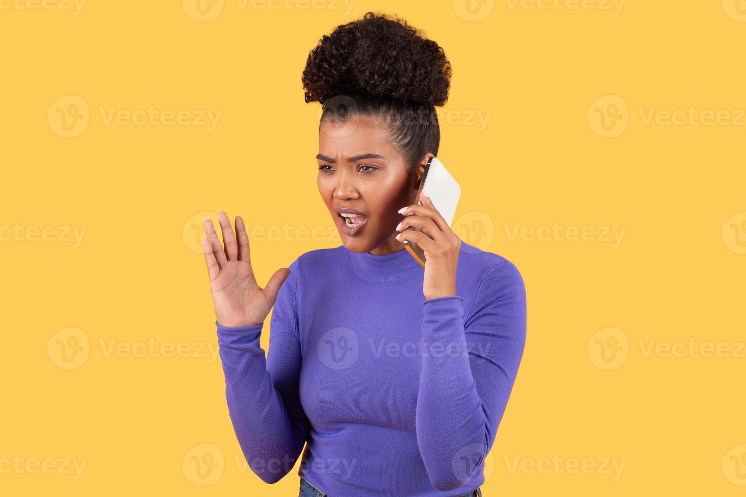 A young Hispanic woman reacts with surprise while speaking on her smartphone. She has curly hair and wears a purple top, set against a bright yellow background, conveying an energetic mood. photo