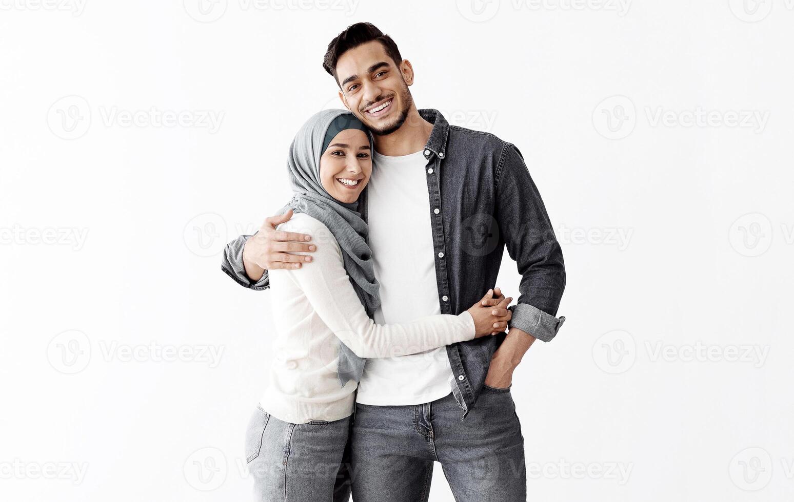 Young muslim couple moving to new apartment, hugging and smiling at camera, standing together in empty room full of paper boxes with their belongings, full length shot, copy space photo