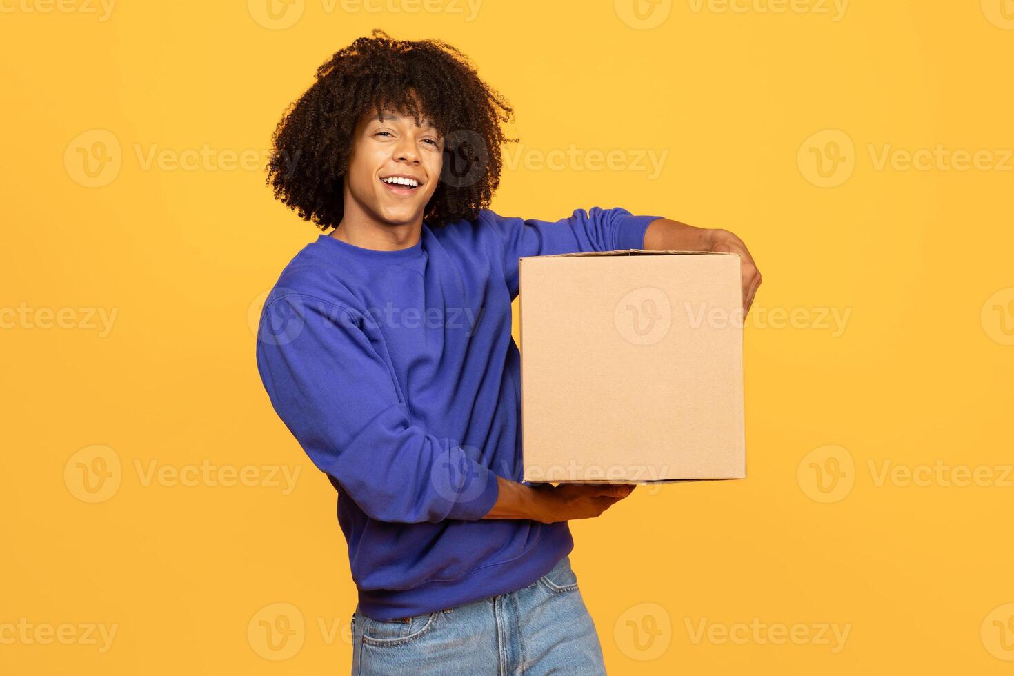 A young black man with curly hair smiles while holding a cardboard box. He stands against a vibrant yellow background, showcasing a cheerful atmosphere perfect for a lifestyle setting. photo