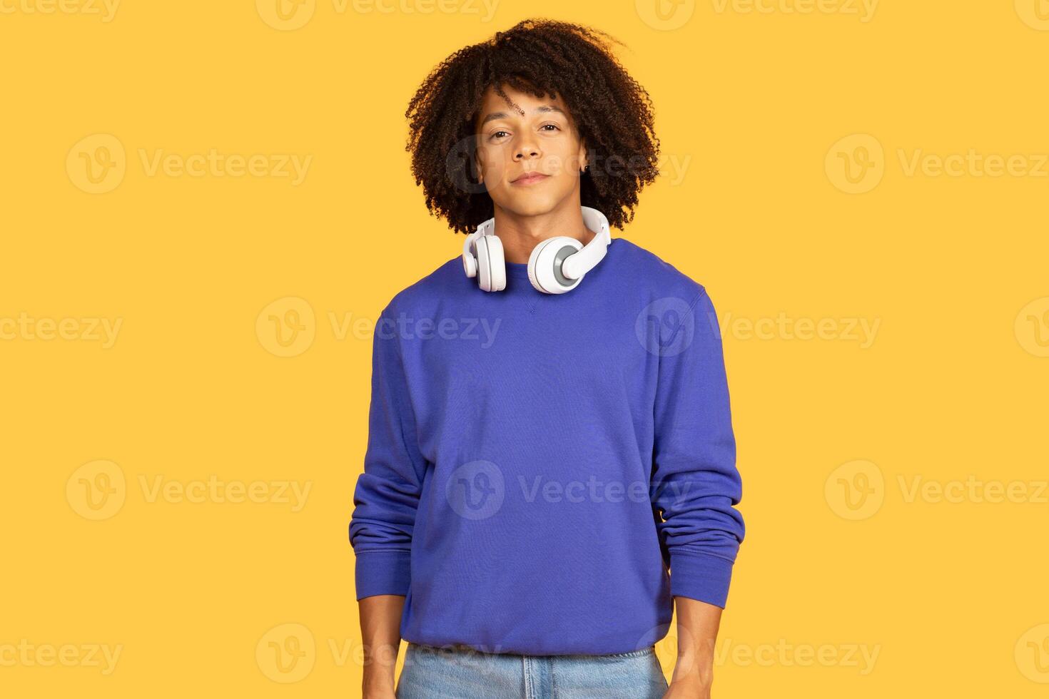 A young black man with curly hair wears headphones while dressed in a blue sweater and jeans. He exhibits a relaxed yet confident attitude against a vibrant yellow background. photo