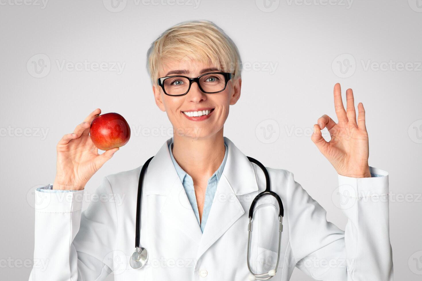 Diet, healthy eating and doctors advice. Smiling female doctor nutritionist in glasses and white coat holds red apple and shows okay sign with hand isolated on blue background, free space, studio shot photo