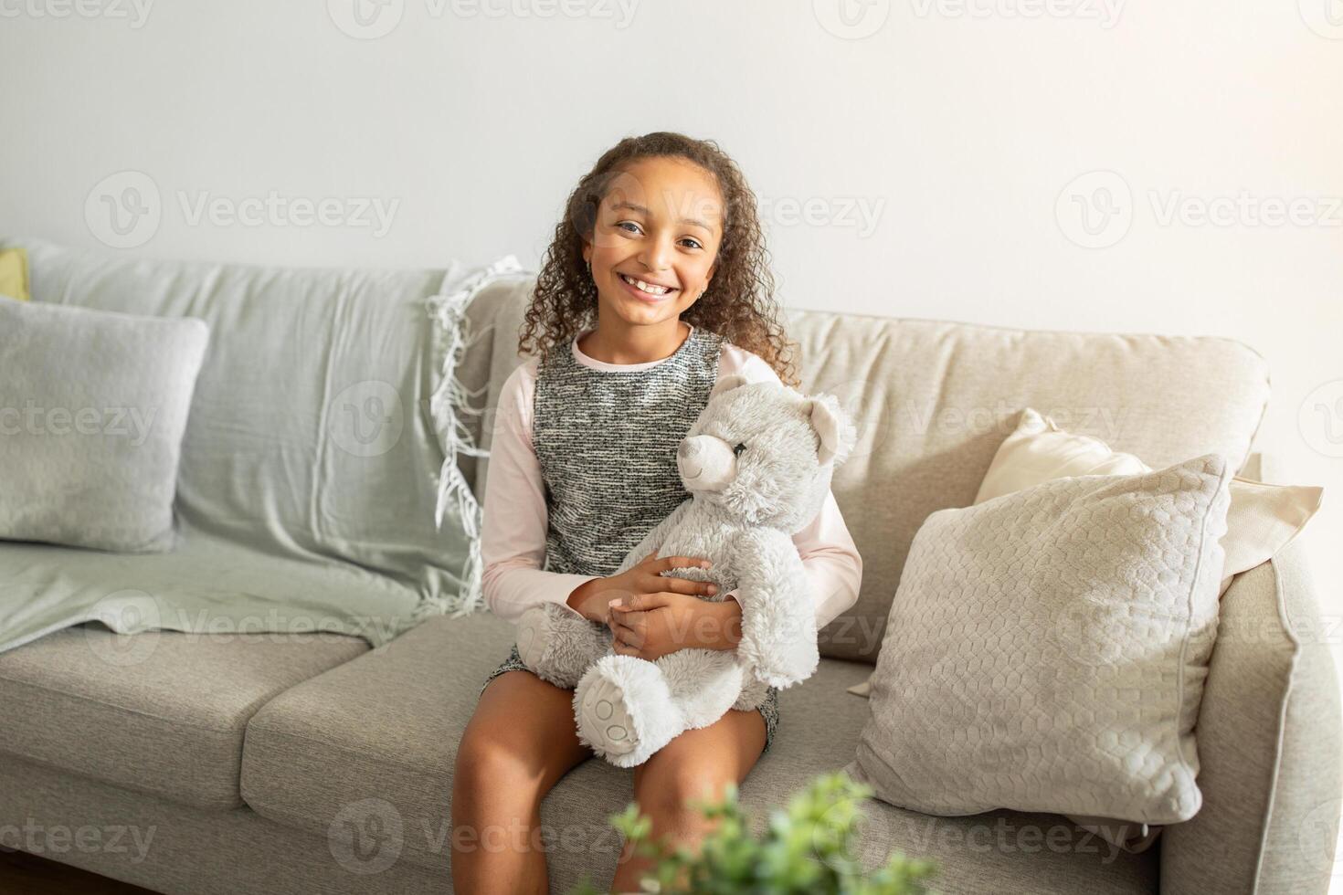 A young African American girl smiles widely while holding a soft teddy bear. She sits comfortably on a cozy couch, surrounded by a peaceful home environment. photo