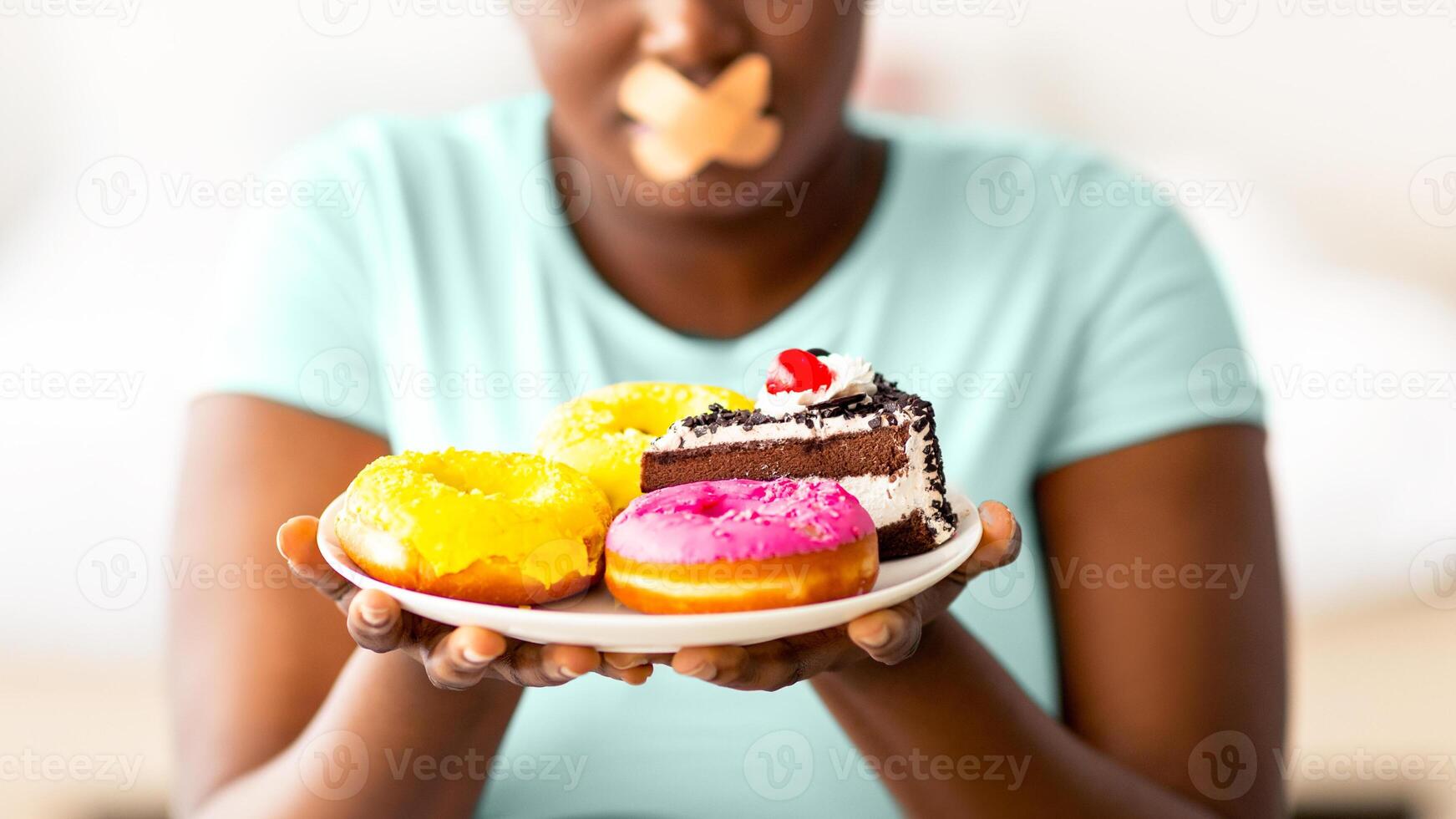 Diet restrictions. Curvy black woman with adhesive bandage on her mouth looking at plate of sweets in hunger at home. Plus size African American lady refusing to eat junk food, losing weight photo