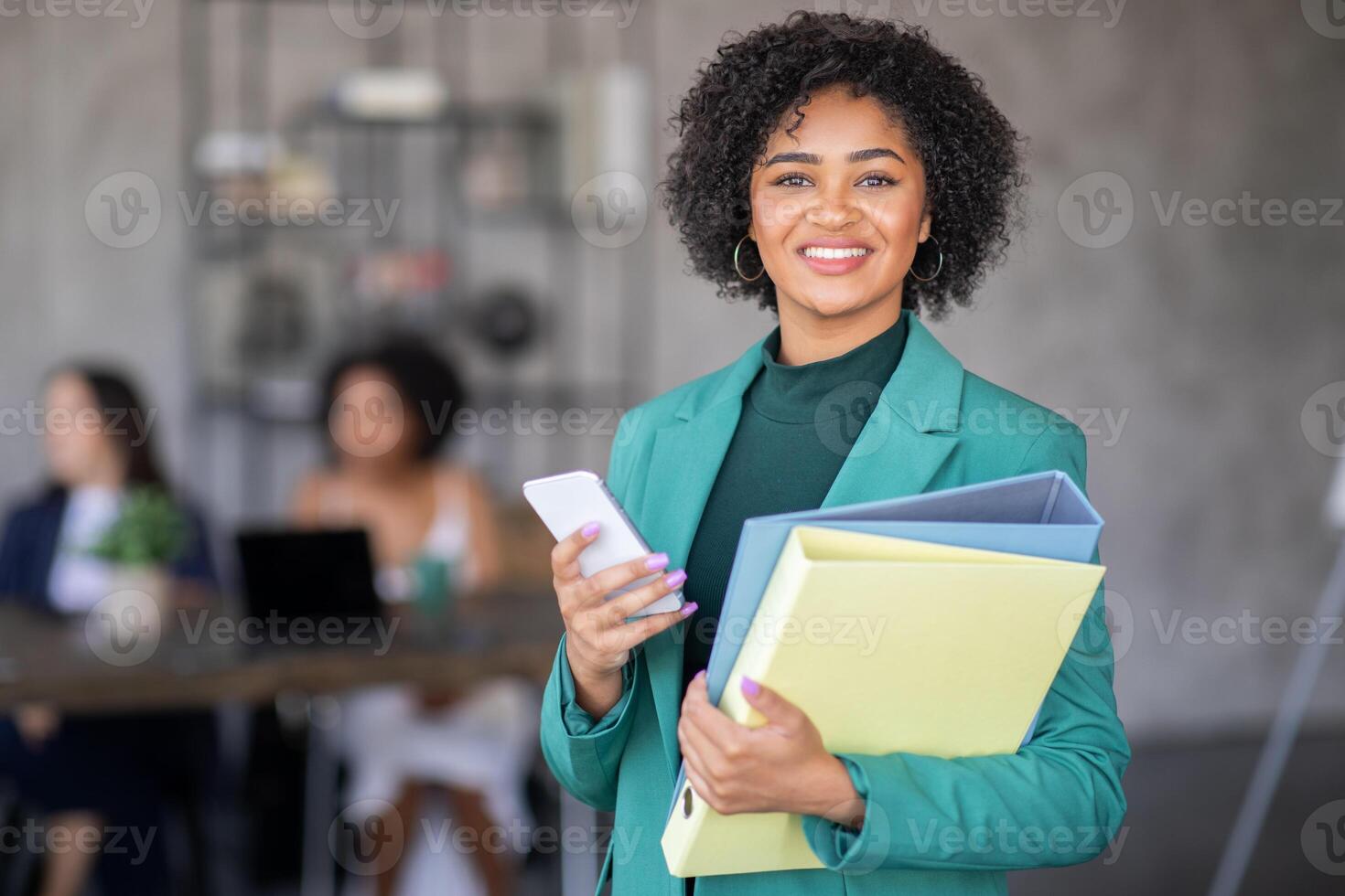 A confident businesswoman stands in a modern office, holding folders and a smartphone. Behind her, colleagues collaborate on a project, emphasizing teamwork and diversity in the workplace. photo