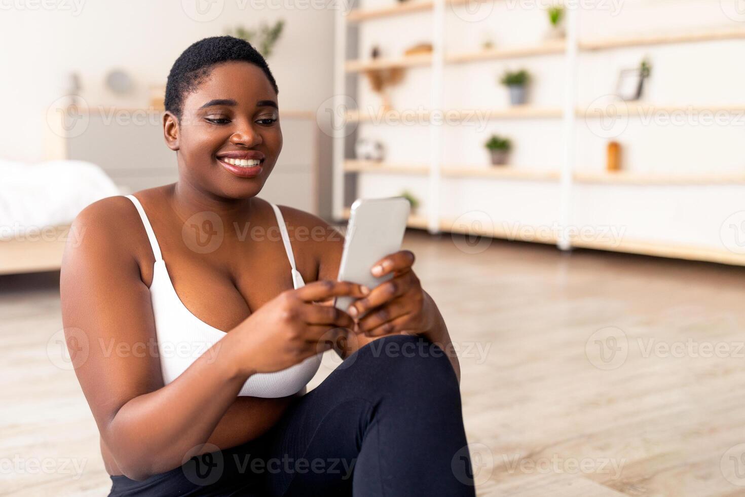 Curvy black lady using mobile device, having break from domestic training, indoors. Plump African American woman browsing social media, resting after home workout photo
