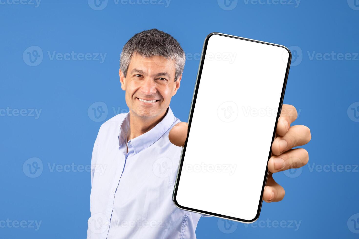 A mature Caucasian man in his 50s smiles while holding a blank smartphone. The bright blue background creates a cheerful, isolated setting that emphasizes his friendly demeanor. photo