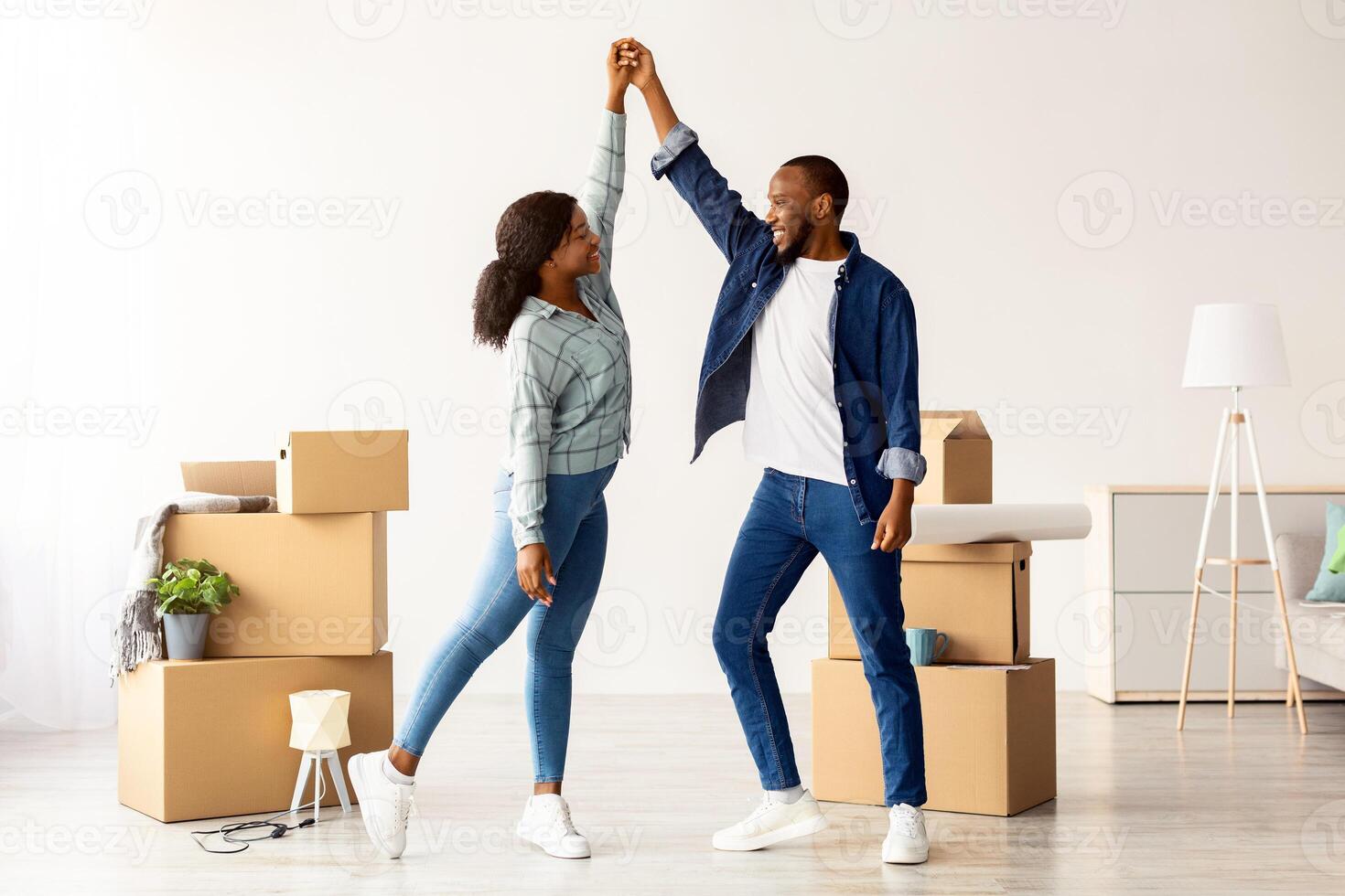 Happy Young Black Couple Celebrating Moving To New Flat, Cheerful African American Spouses Dancing Together In Bright Room Among Unpacked Cardboard Boxes With Belongings, Recolation Concept photo