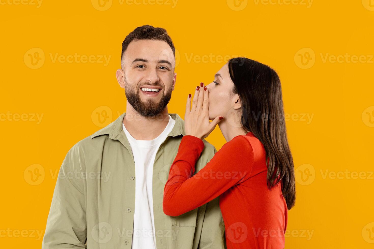 A couple stands close together, with the woman whispering sweetly into the man's ear. Their smiles reflect joy and intimacy, highlighting their bond in a lively setting. photo
