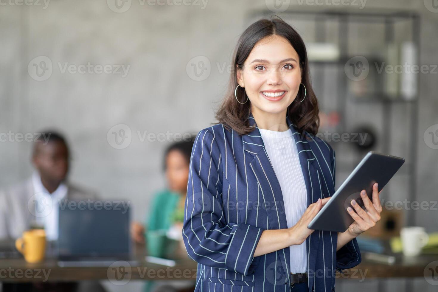 A confident woman stands in front of her diverse team in a contemporary office. She holds a tablet and smiles, as colleagues engage in a productive discussion while working together on projects. photo