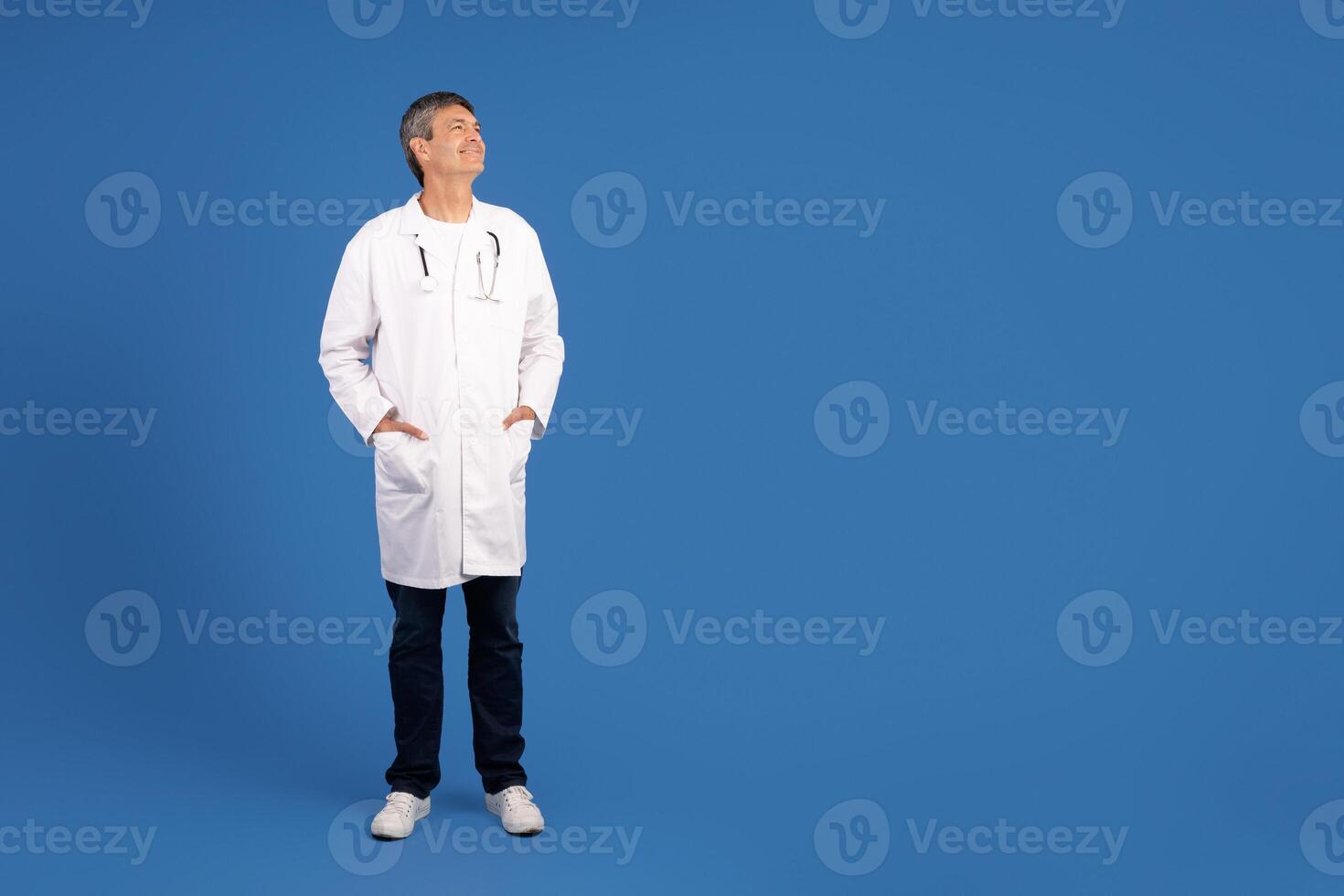 A middle aged European man, dressed in a lab coat, stands with hands in pockets and gazes upward. The confident doctor embodies a professional healthcare attitude in a minimalist setting, copy space photo
