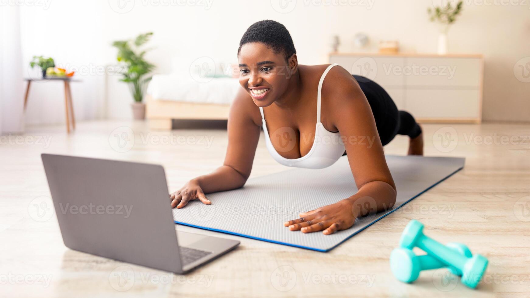 Plump black lady exercising at home, following her personal trainer's online instructions on laptop, standing in plank pose. Plus size African American woman working out to tutorial on web photo