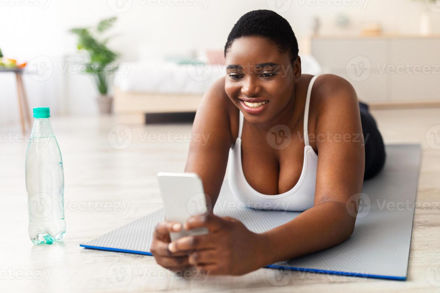Happy plus size black woman using mobile phone on her break from domestic training, browsing web, looking up workout online. Overweight African American lady checking cellphone after exercising photo
