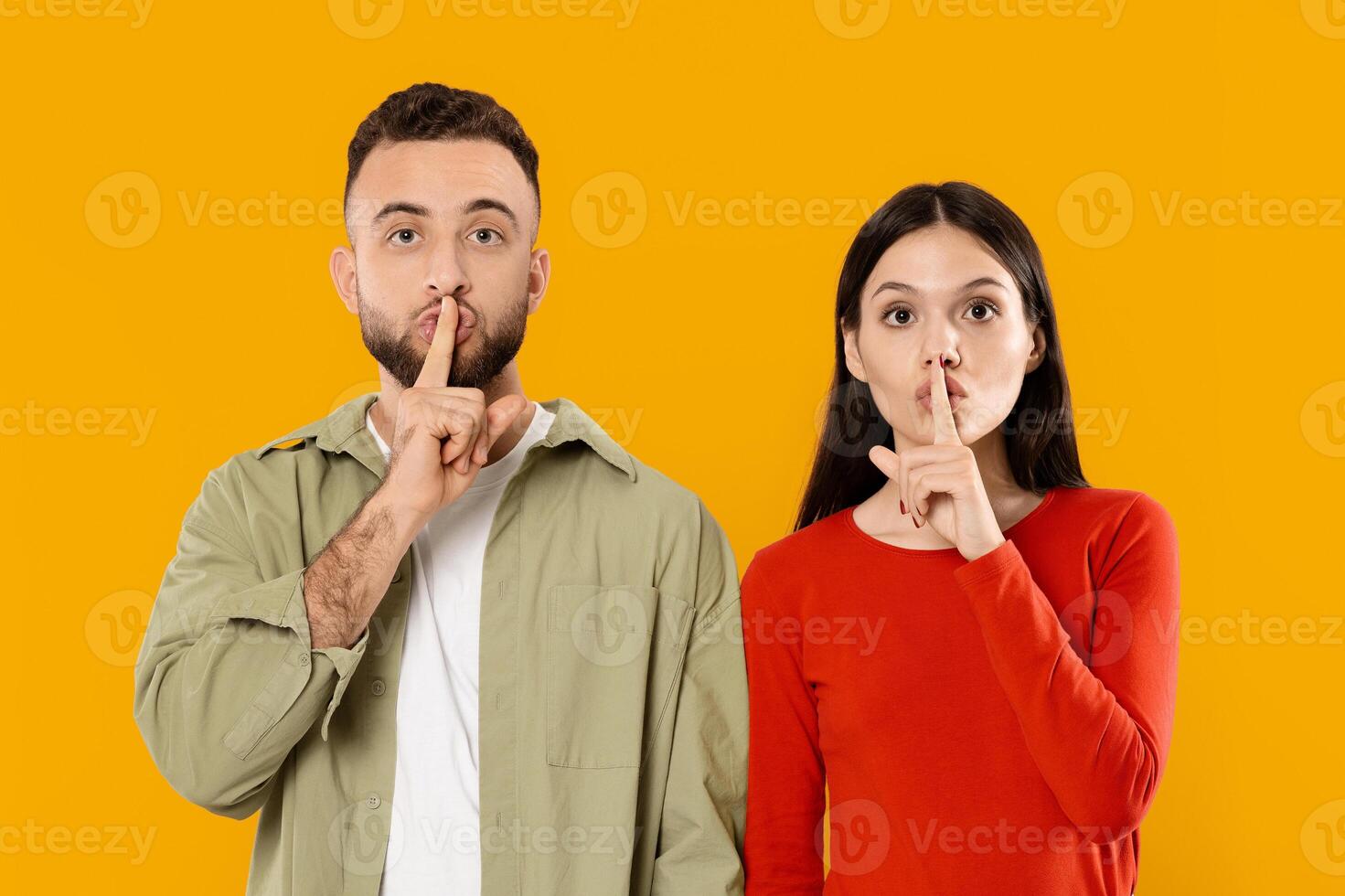 A young married couple poses together with their fingers to their lips, expressing a playful gesture of secrecy or silence against a bright yellow backdrop, showcasing their affection. photo