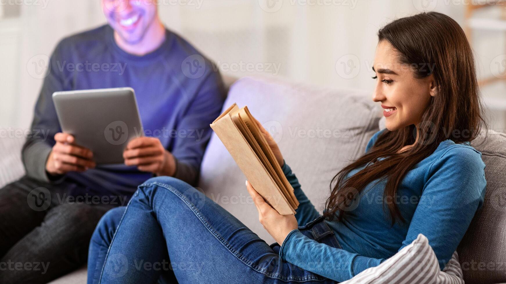 Free Time Concept. Happy spouses sitting on couch in their apartment, girl reading book, boy using tablet photo