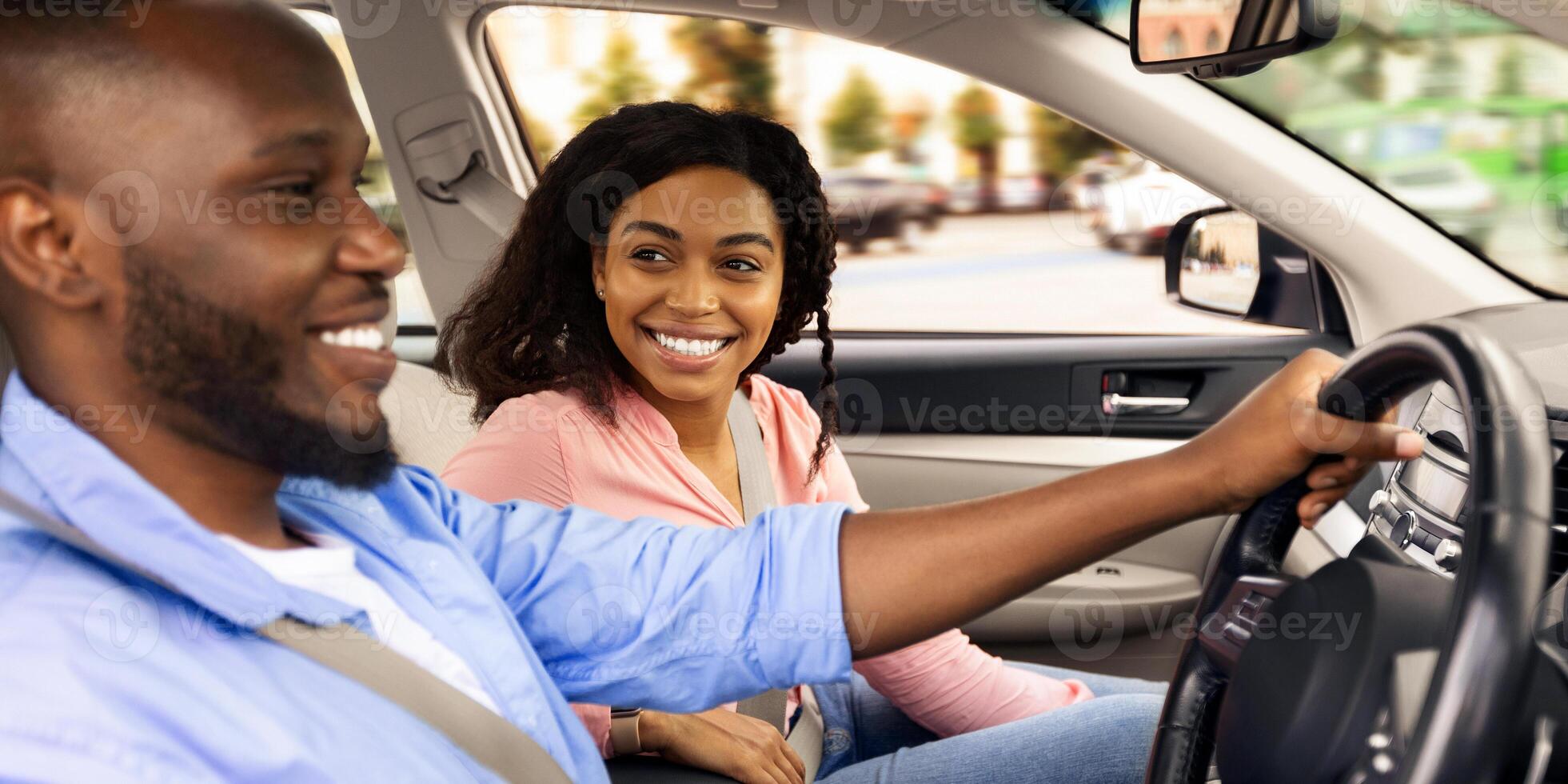Enjoying Travel Concept. Smiling African American man driving modern luxury car, happy young woman sitting on the front passenger seat, looking on the driver, family going on roadtrip, side view photo