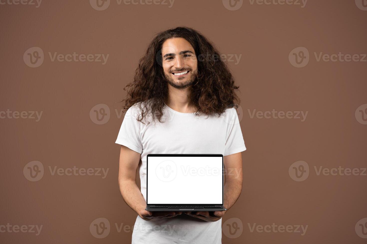 A young European man with long, curly hair stands against a plain brown backdrop, smiling while holding a laptop with a blank screen. He embodies a modern, lifestyle-oriented vibe. photo