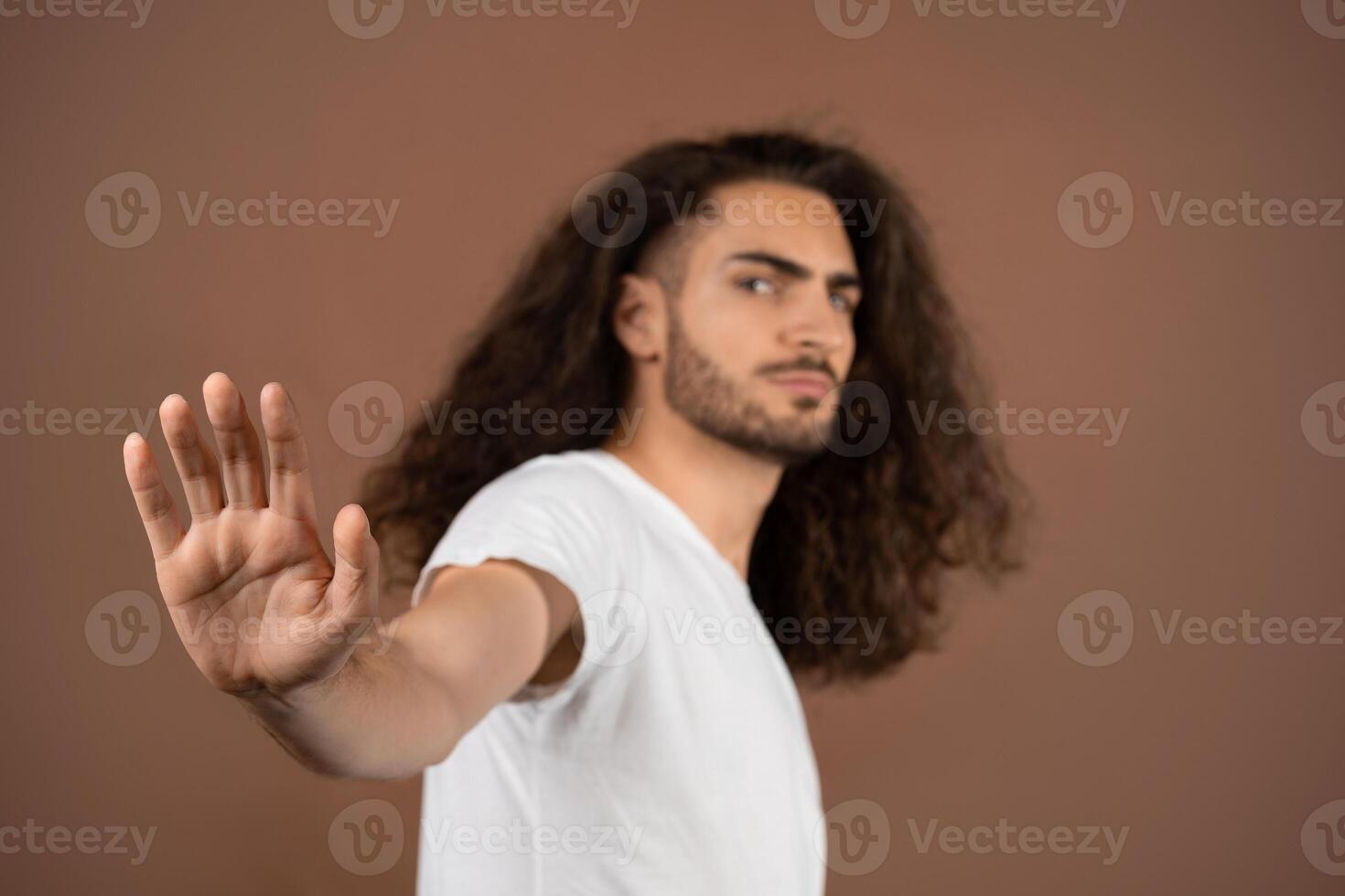 A young European man with long, curly hair stands in a relaxed pose, raising one hand as if signaling to stop. He wears a simple white t-shirt and showcases a thoughtful expression. photo