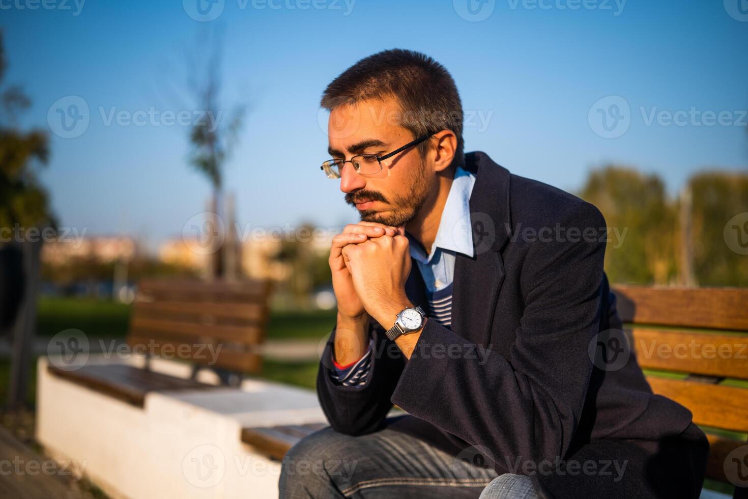 Worried businessman in coat with eyeglasses sitting on the bench in the park on a sunny autumn day. photo