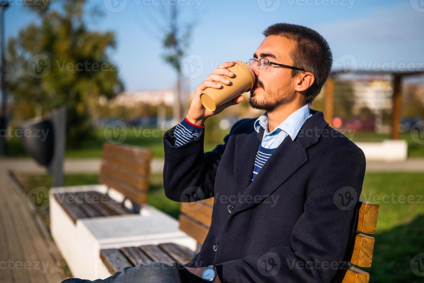 Businessman in coat with eyeglasses drinking coffee on the bench in the park on a sunny autumn day. photo