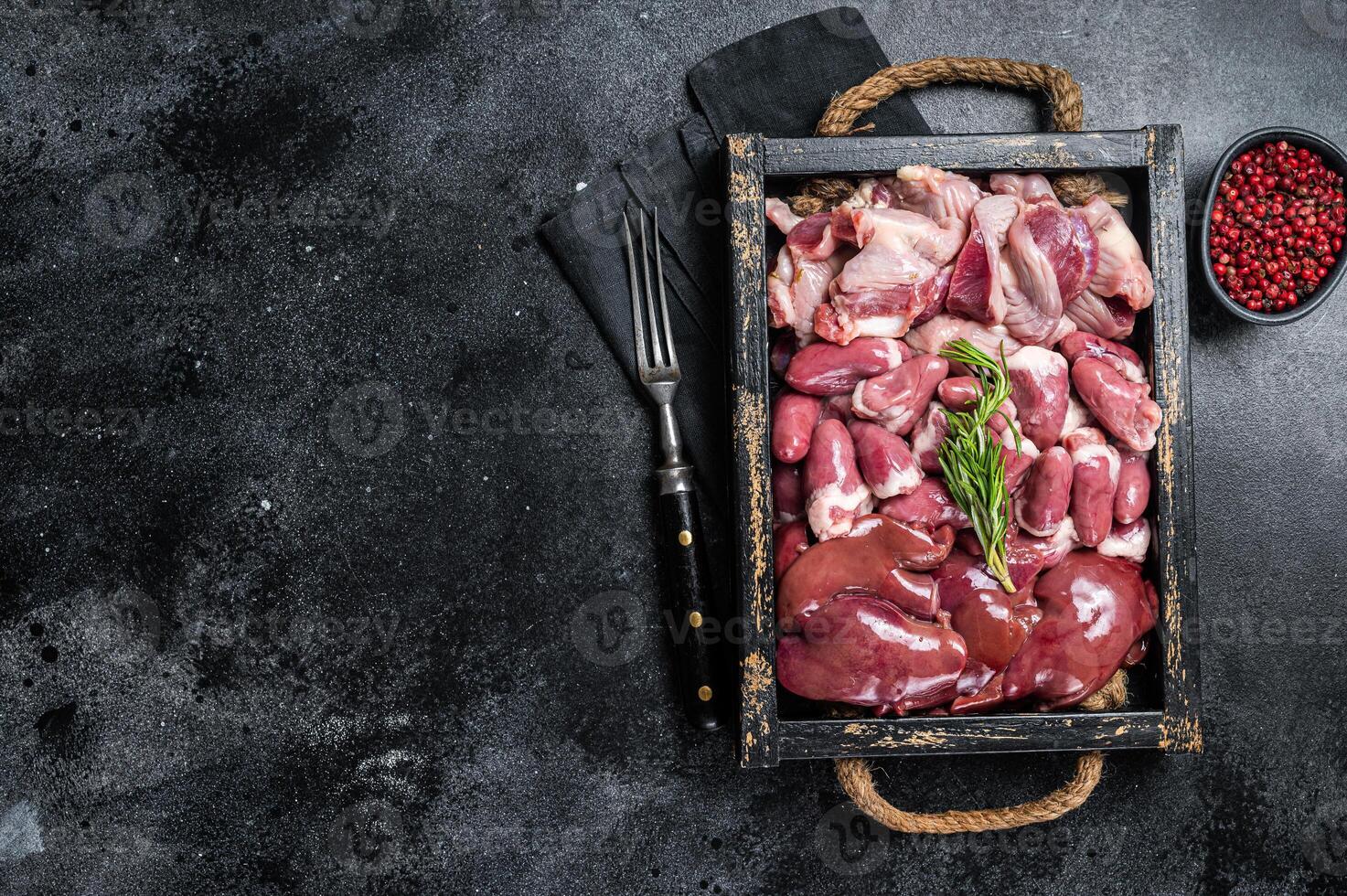 Uncooked Bird offals, raw chicken giblets, gizzards, stomachs, liver and hearts in a wooden tray. Black background. Top view. Copy space photo