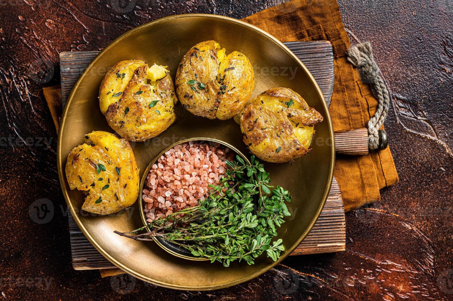 Crushed, smashed potatoes baked with rosemary and thyme on plate. Dark background. Top view photo