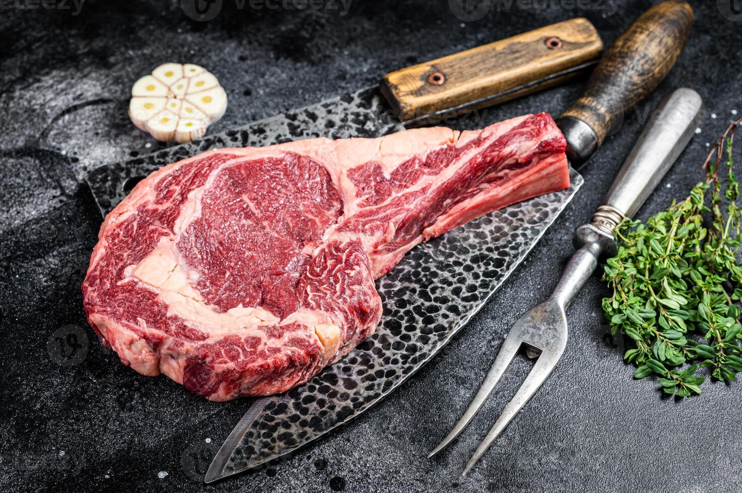 Dry aged raw tomahawk or cowboy beef steak on a black table with knife, cleaver, spice and herbs. Black background. Top view photo
