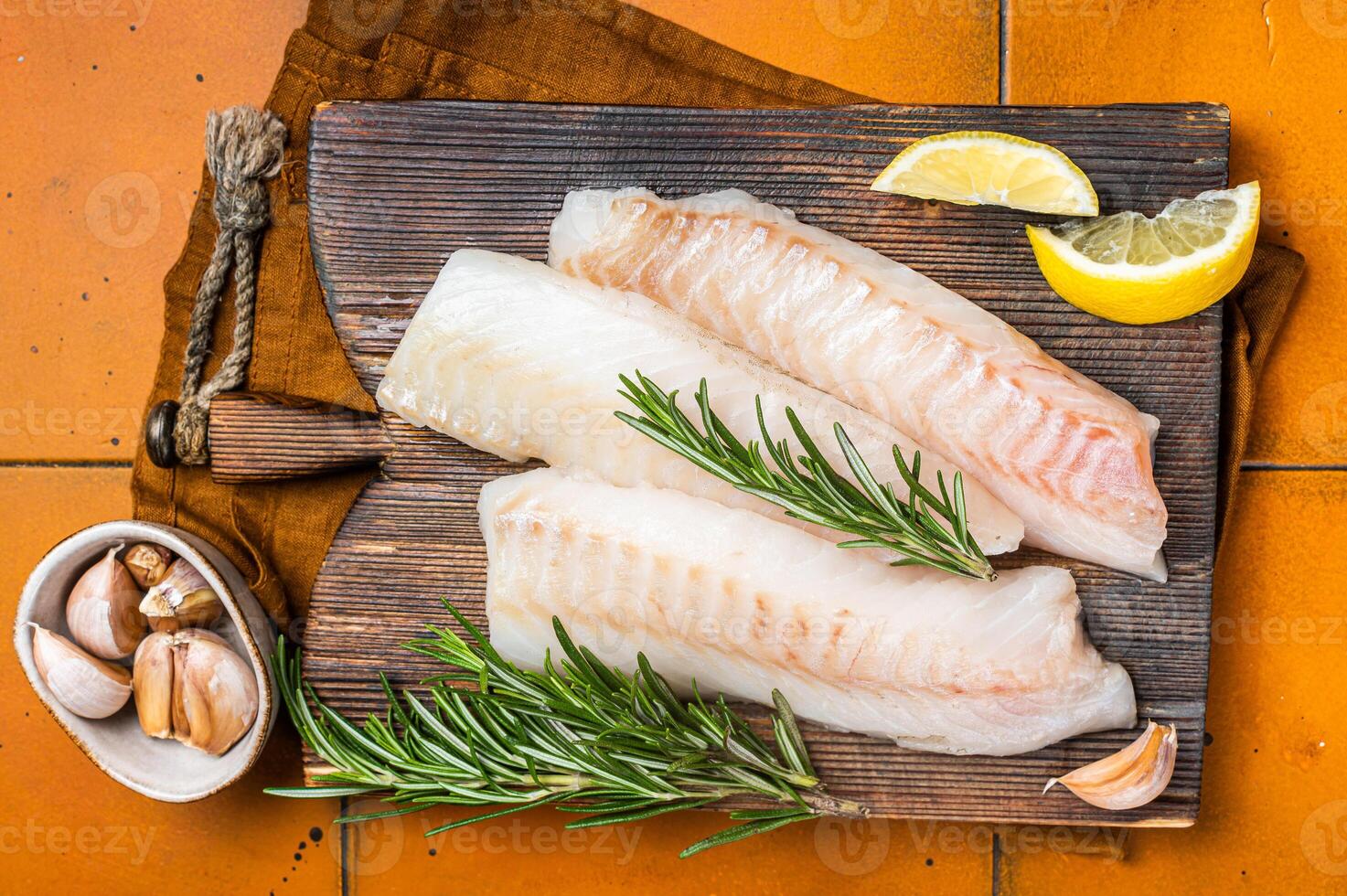 Raw cod fish fillets, codfish with rosemary on wooden board. Orange background. Top view photo