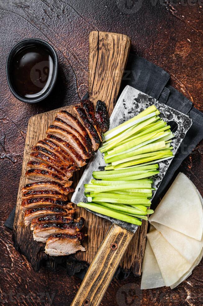 Sliced Peking Duck served with cucumber, green onion, and wheaten chinese pancakes on a meat cleaver. Dark background. Top view photo