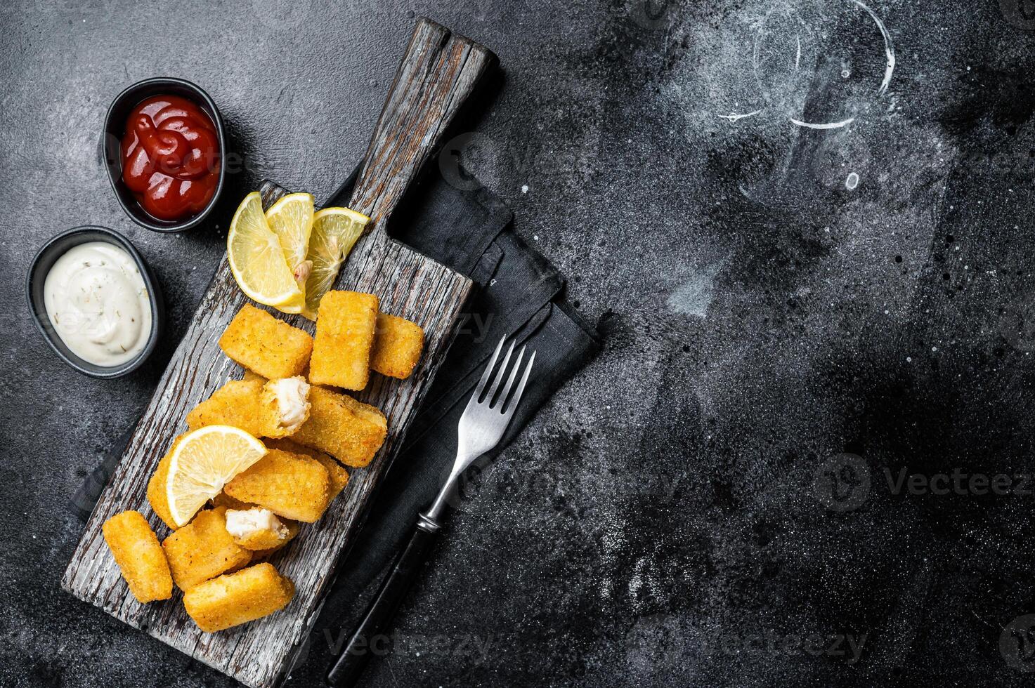 Golden roast Fish Sticks Fingers with sauce on a wooden board. Black background. Top view. Copy space photo