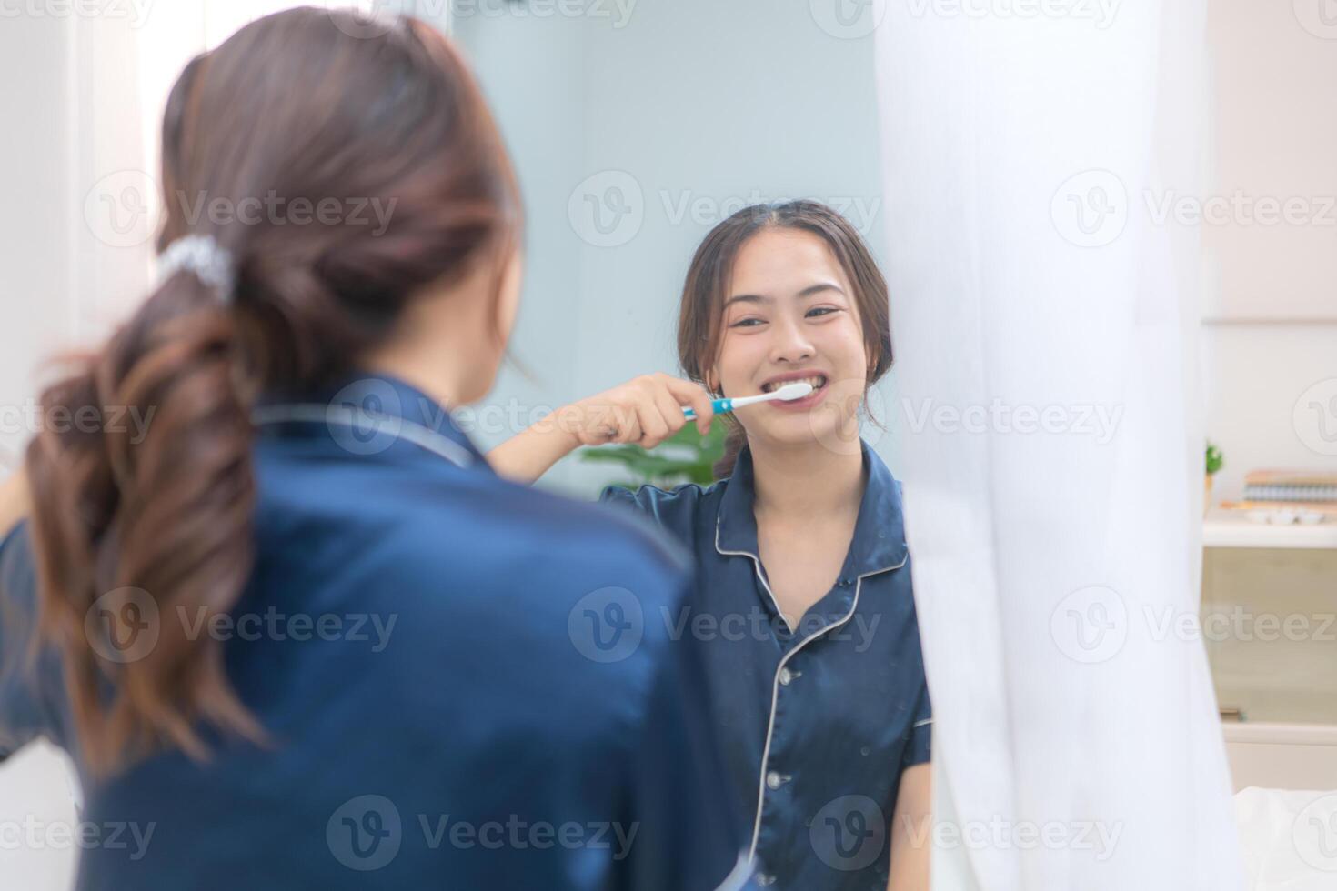 Asian young woman in a blue pajama is brushing her teeth in front of the mirror before taking a shower photo