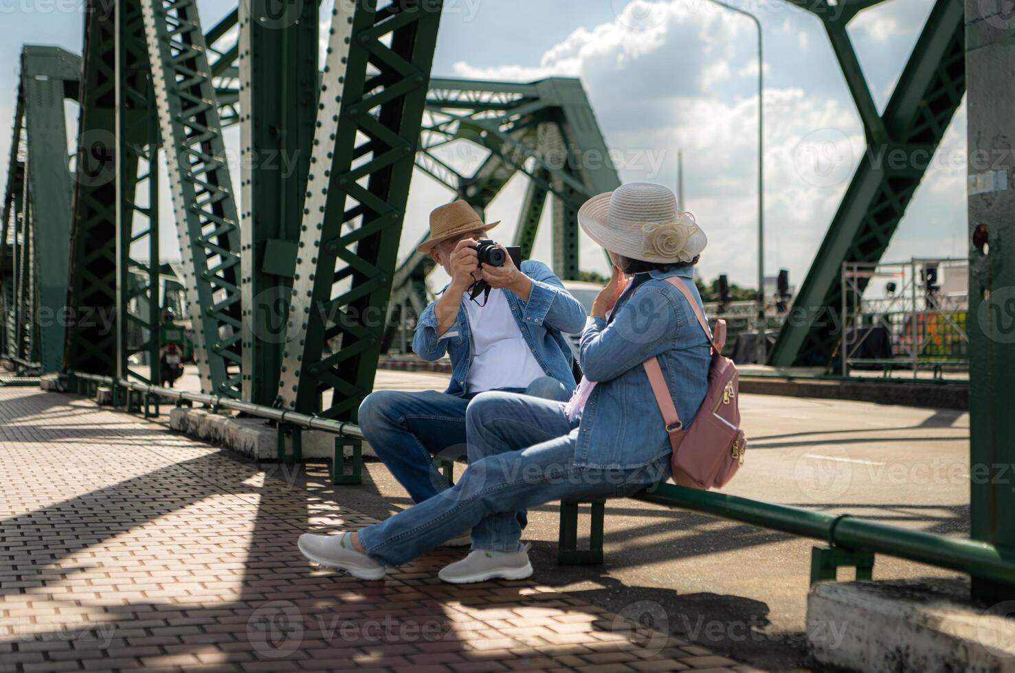 An elderly couple relaxes and looks at photos in camera after walking through the city streets with the road and bridge structures in the background