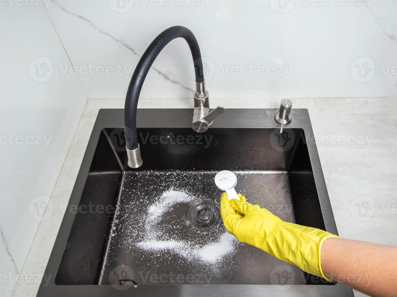 A man's hand in a yellow glove holds a spoon with oxygen bleach against the background of a kitchen sink. Cleaning from grease and dirt. photo