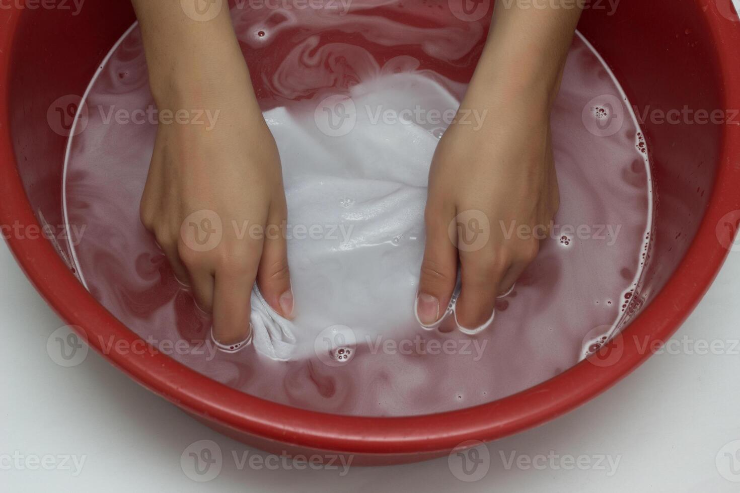 The girl erases a children's white T-shirt in a red basin from stains, cleans from stain remover, close-up, hands, white background photo