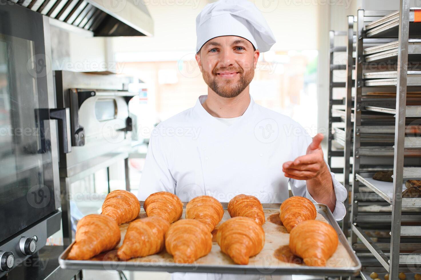 Handsome baker in uniform holding tray full of freshly baked croissants at the manufacturing photo
