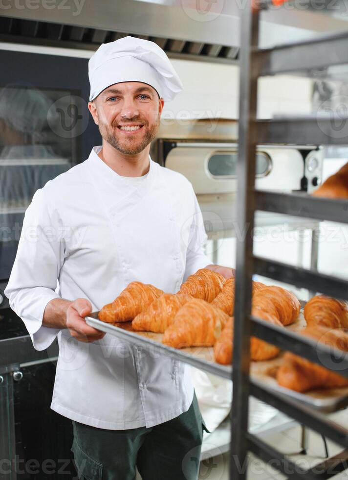 A young handsome male baker holds a tray with French croissants in front of a bakery and smiles photo
